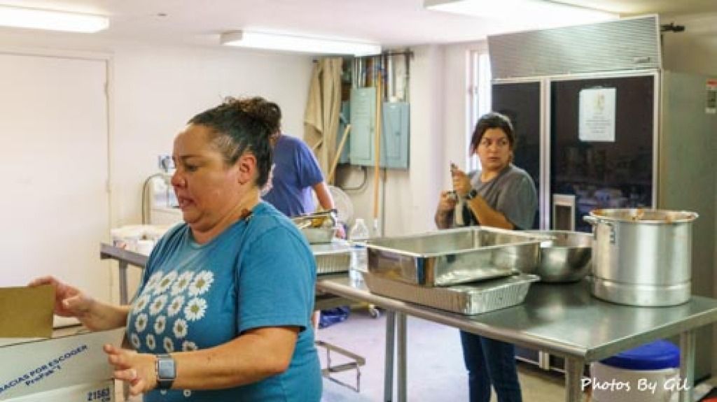 A woman in a blue daisy shirt packs a box in a kitchen with stainless steel counters. 