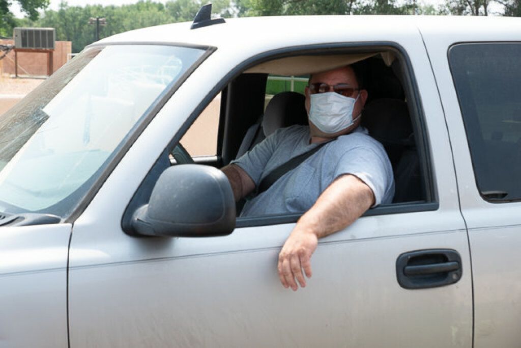 Man in a gray T-shirt and mask drives a white pickup truck, arm resting on the open window.