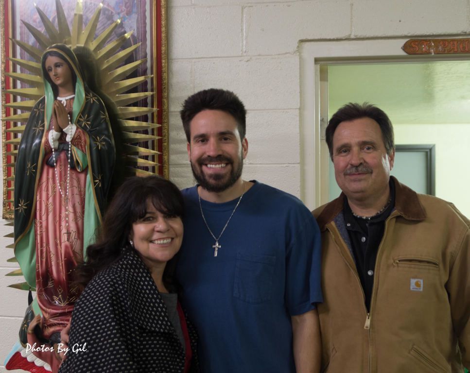 A smiling group of three people, two men and a woman, stand indoors next to a statue of the Virgin Mary. 