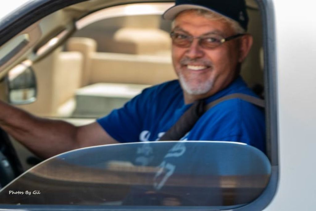 Smiling man in glasses and cap, sitting in driver's seat of a vehicle, wearing a blue shirt. 