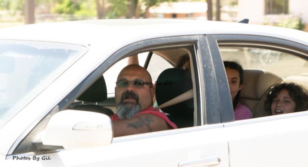 A man with glasses drives a white car, with children in the back seat looking out.