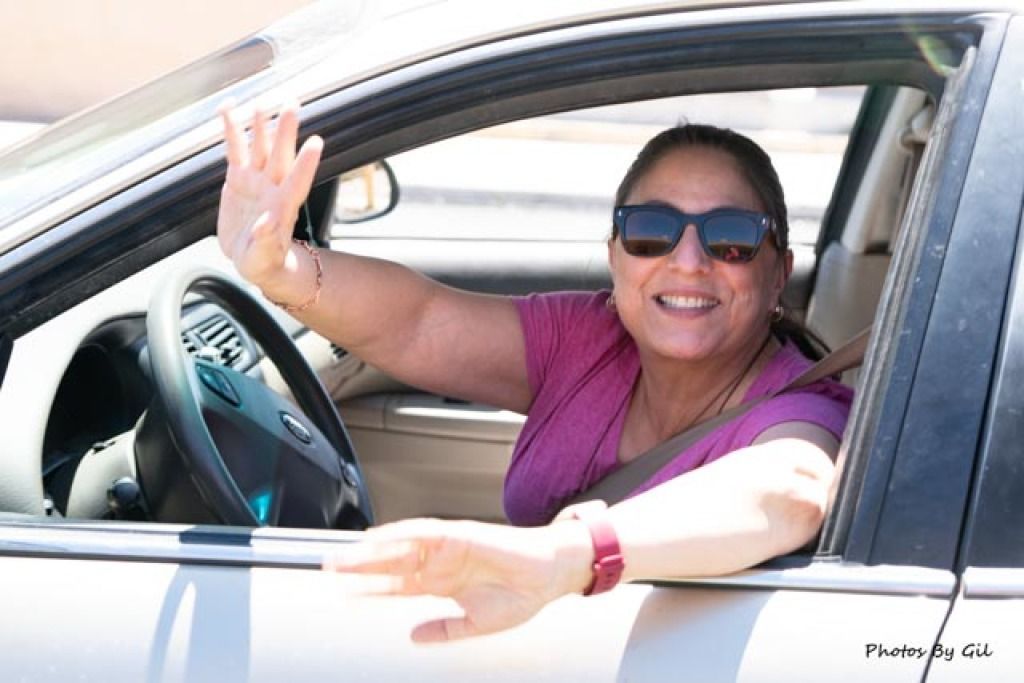 A woman wearing sunglasses and a pink shirt smiles and waves from a car window.