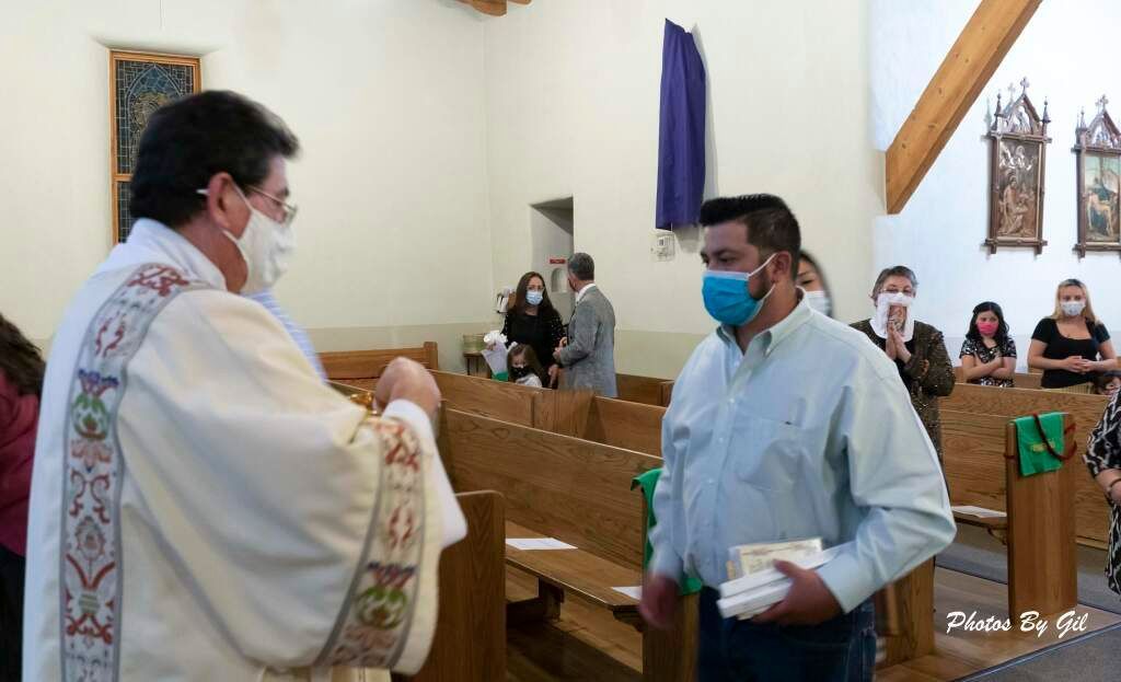 A priest in a decorated robe, wearing a mask, hands a sacramental item to a masked man in a light blue shirt inside a church.