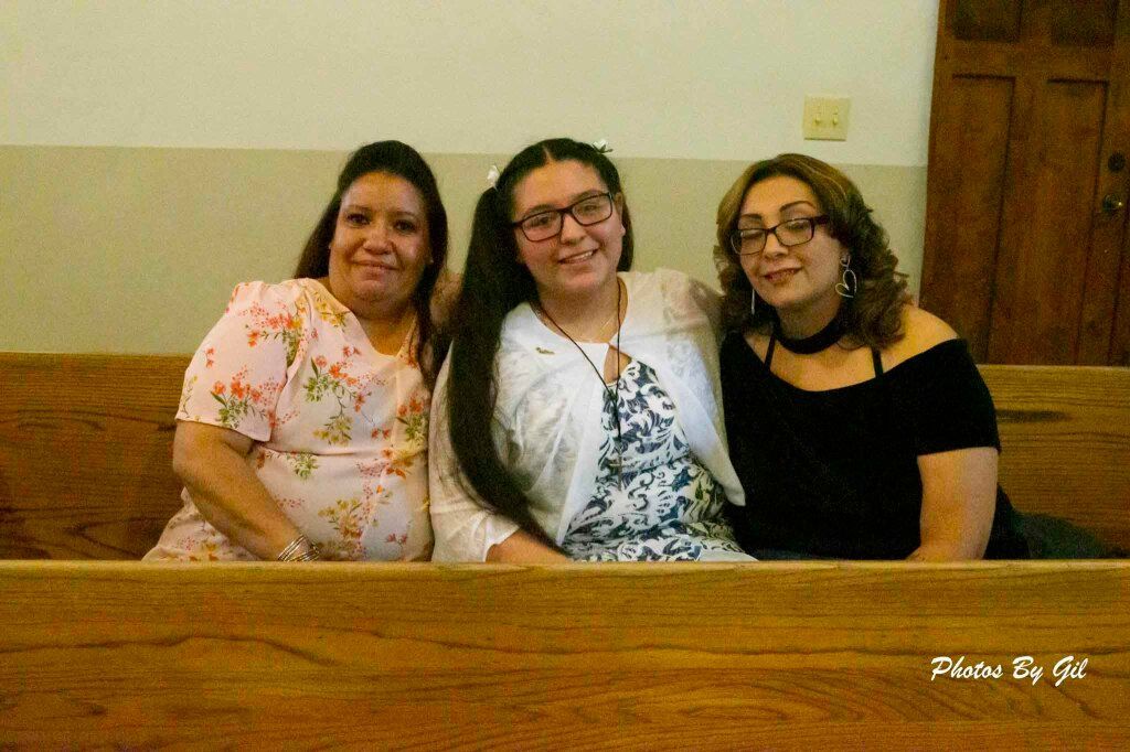 Three women sit closely on a wooden pew, smiling warmly at the camera. 