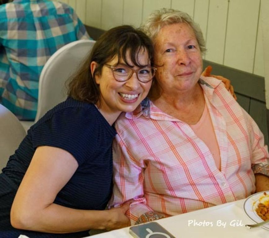 A young woman with glasses and dark hair smiles brightly, hugging an older woman in a pink plaid shirt.