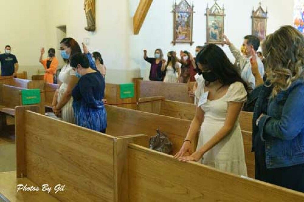 People in a church, wearing face masks and standing in wooden pews, bow their heads during a ceremony.