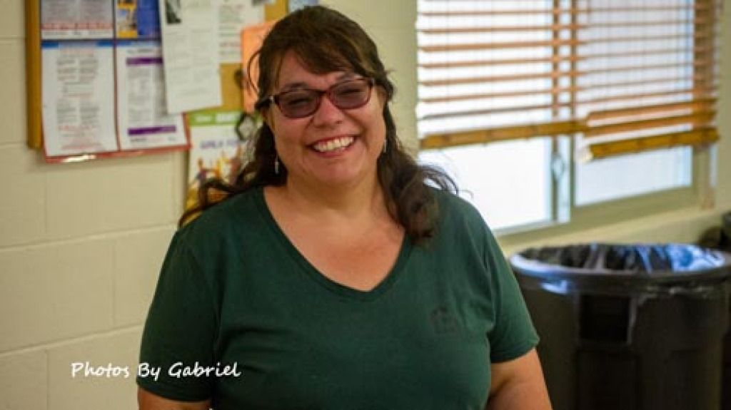 A woman with long brown hair and glasses smiles warmly, wearing a green shirt. 