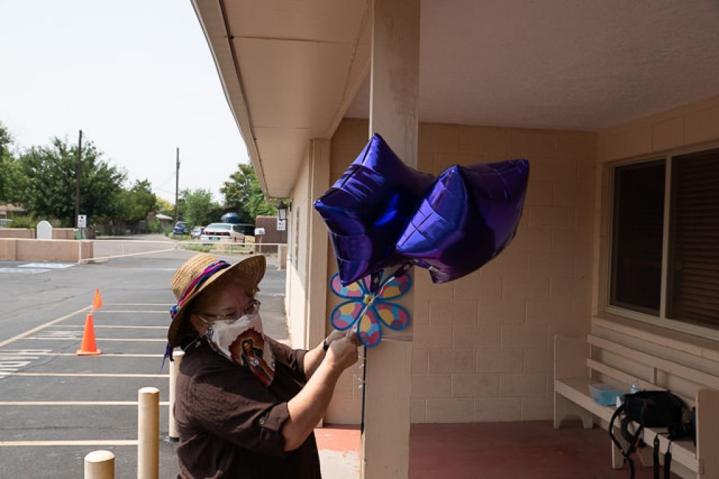 A woman in a straw hat and face mask ties star-shaped purple balloons to a post outside a building. 