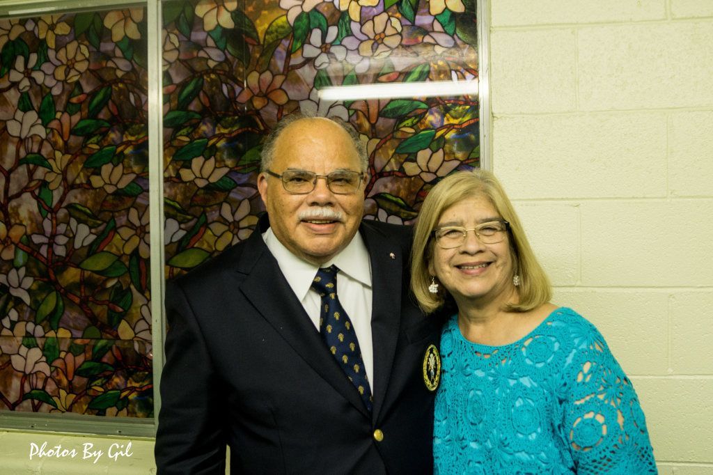 An elderly couple smiles warmly, standing in front of a vibrant stained glass window with floral motifs.