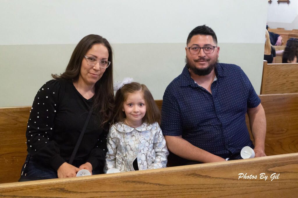 A family of three, a woman, a child, and a man, sit in a wooden pew, smiling warmly.