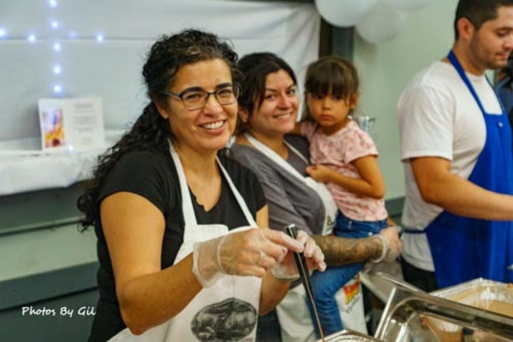 Smiling woman with glasses and apron serves food in a lively, community kitchen.
