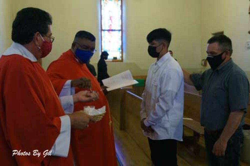 A priest and religious leader in red robes perform a ceremony for a young man in a white shirt.