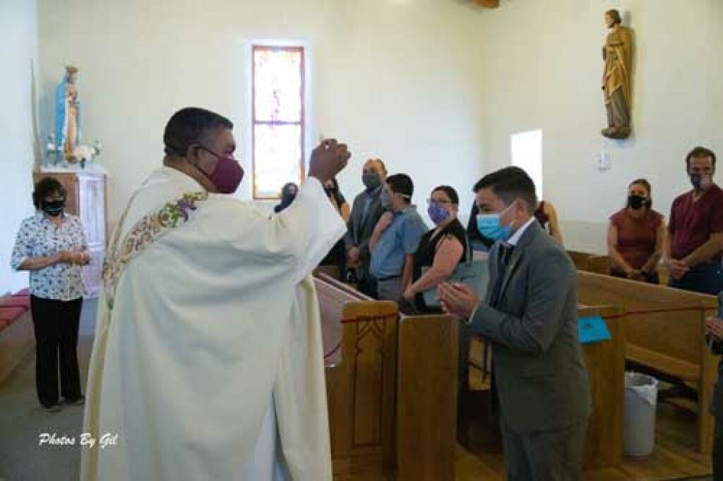 A priest in white robes, wearing a mask, performs a blessing for a masked man in a suit in a church.