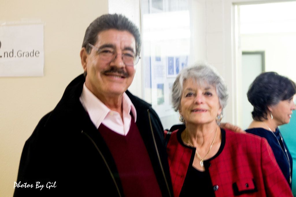 A man and woman smiling warmly in a hallway, both wearing red and black attire. 