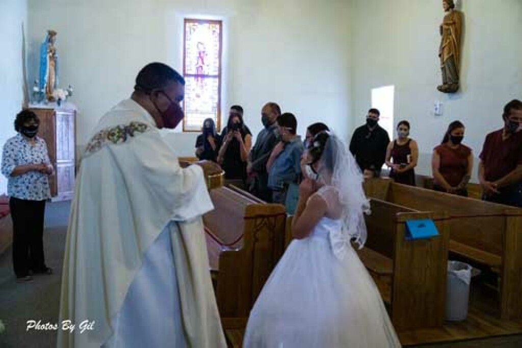 A priest in a white robe and red mask conducts a ceremony with a young girl in a white dress and veil. 