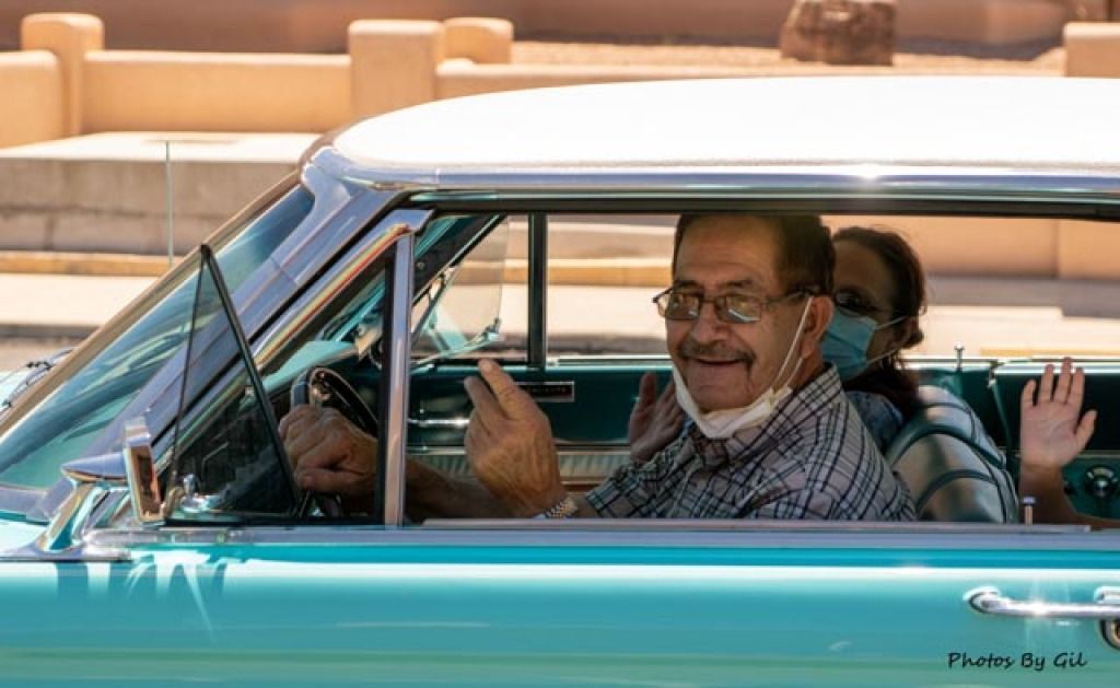 A smiling man wearing glasses and a mask pulls down in a vintage teal car.