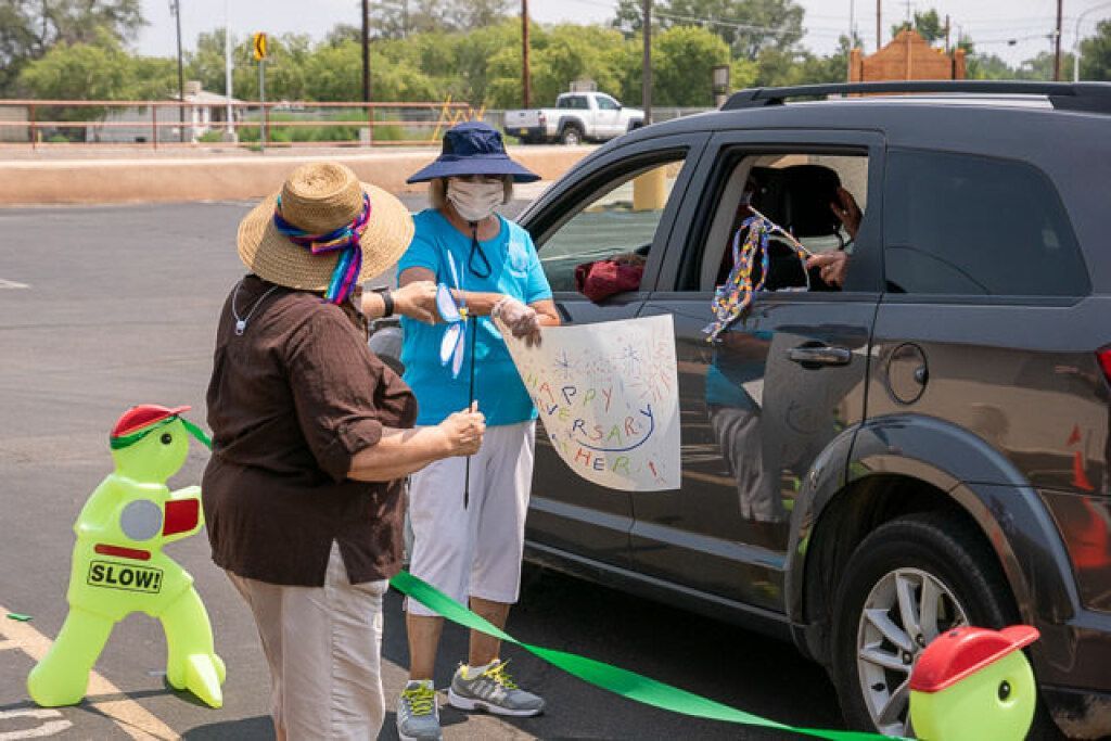 Two women in hats and masks celebrate beside a car in a parking lot. 