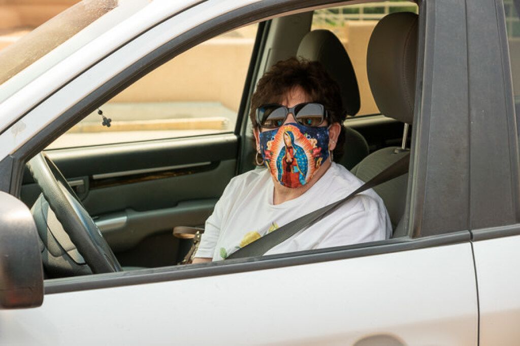 A person wearing sunglasses and a colorful mask with a religious image sits in the driver's seat of a white car.