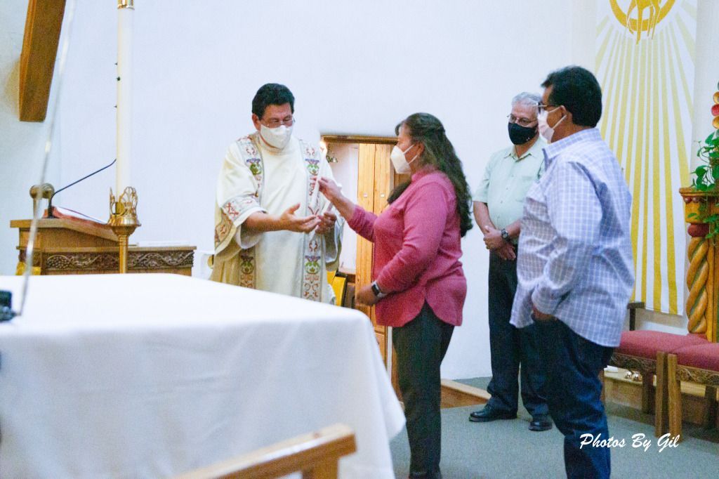 A priest in white robes hands a lit candle to a masked woman in a church ceremony.