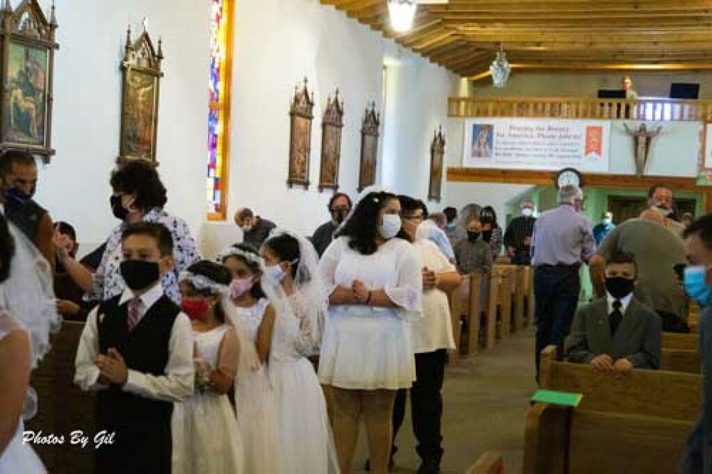 Children in white dresses and suits, wearing face masks, are lined up for a ceremony inside a decorated church.