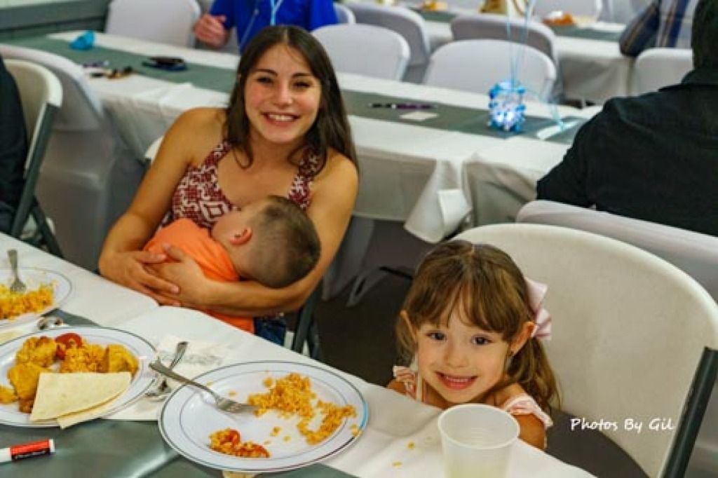 A woman smiling while holding a sleeping baby at a table with food. 