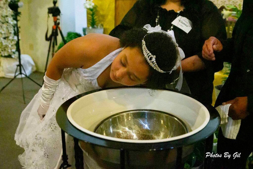A young girl in a white dress and tiara leans over a baptismal font, preparing for a baptism ceremony. 