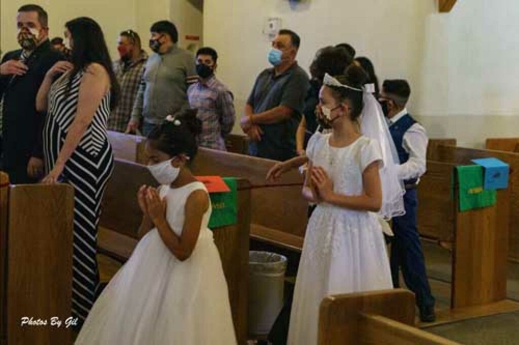 Young girls in white dresses, possibly for a religious ceremony, stand in a church aisle, hands clasped.