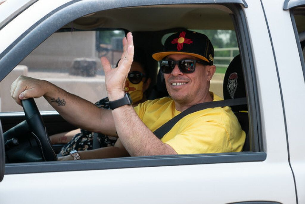 A man in sunglasses and a black cap waves from a white car, smiling. 