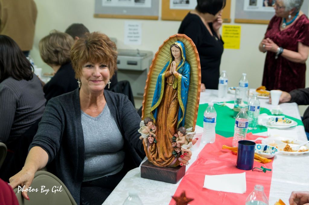 A woman smiles at a community gathering, sitting beside a colorful statue of the Virgin Mary. 