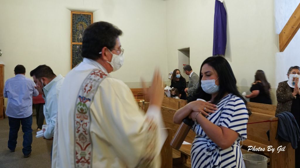 A priest in a decorated robe, wearing a mask, blesses a woman in a striped dress in a church setting. 
