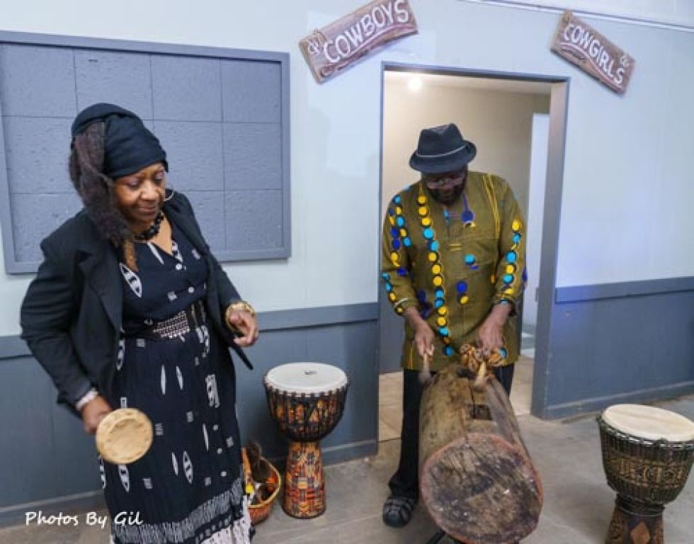 Two people play drums indoors, surrounded by various percussion instruments. 