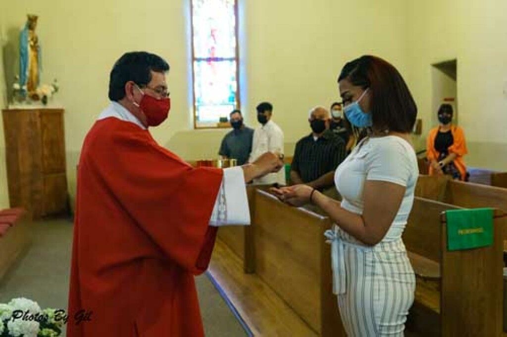 A priest in red vestments gives communion to a masked woman in a church. 