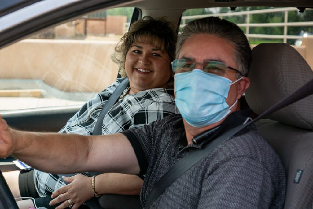 A man wearing a mask and a woman smiling sit in a parked car.