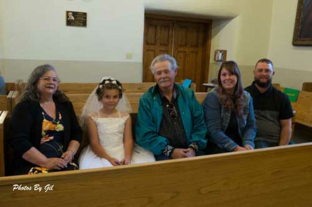 A young girl in a white dress and veil sits on a church pew with adults.