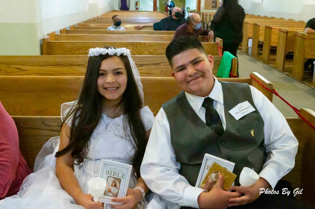 A young girl and boy sit smiling in a church pew, dressed in formal attire.