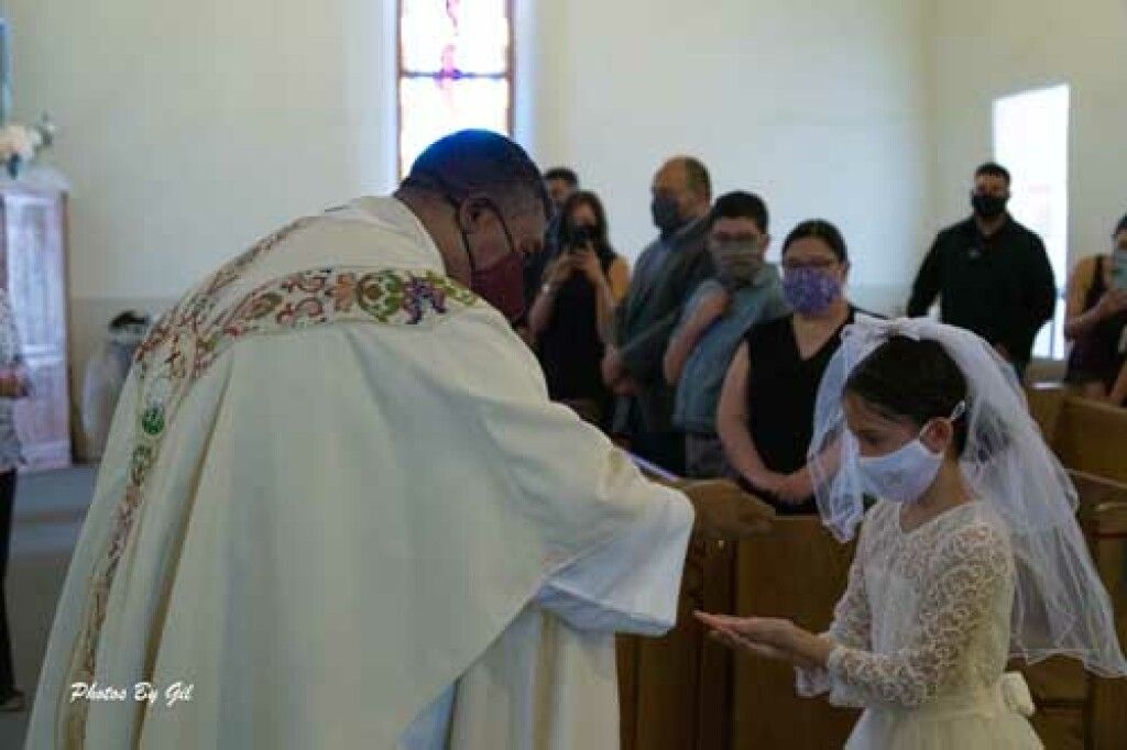 A priest in a decorated white robe offers communion to a girl in a white dress and veil. 