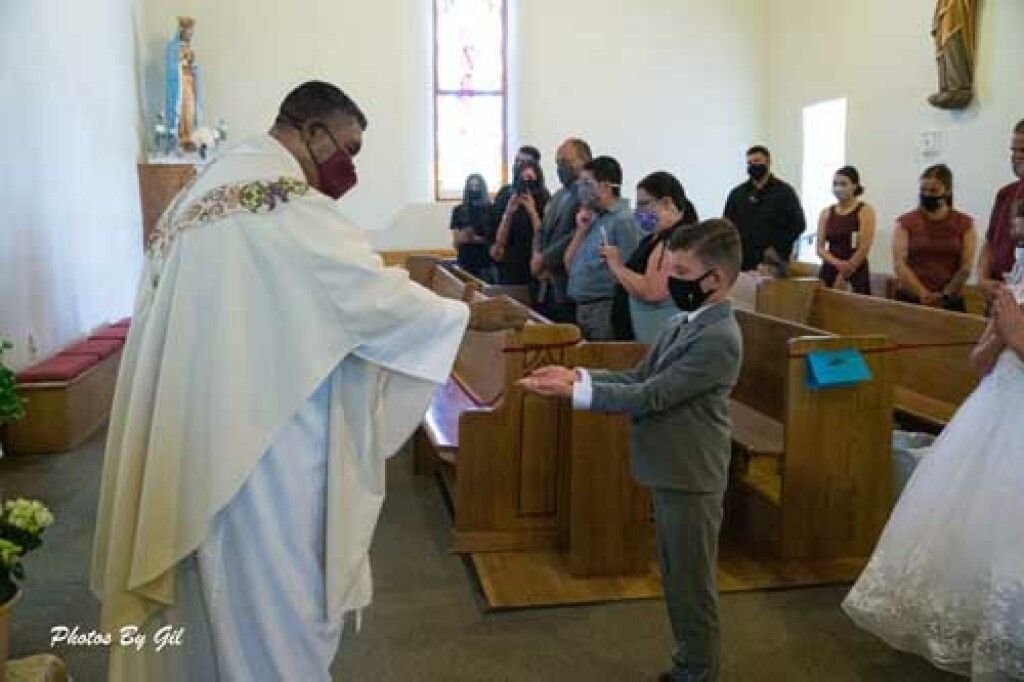 A priest in a white robe and red mask gives communion to a boy in a suit at a church.