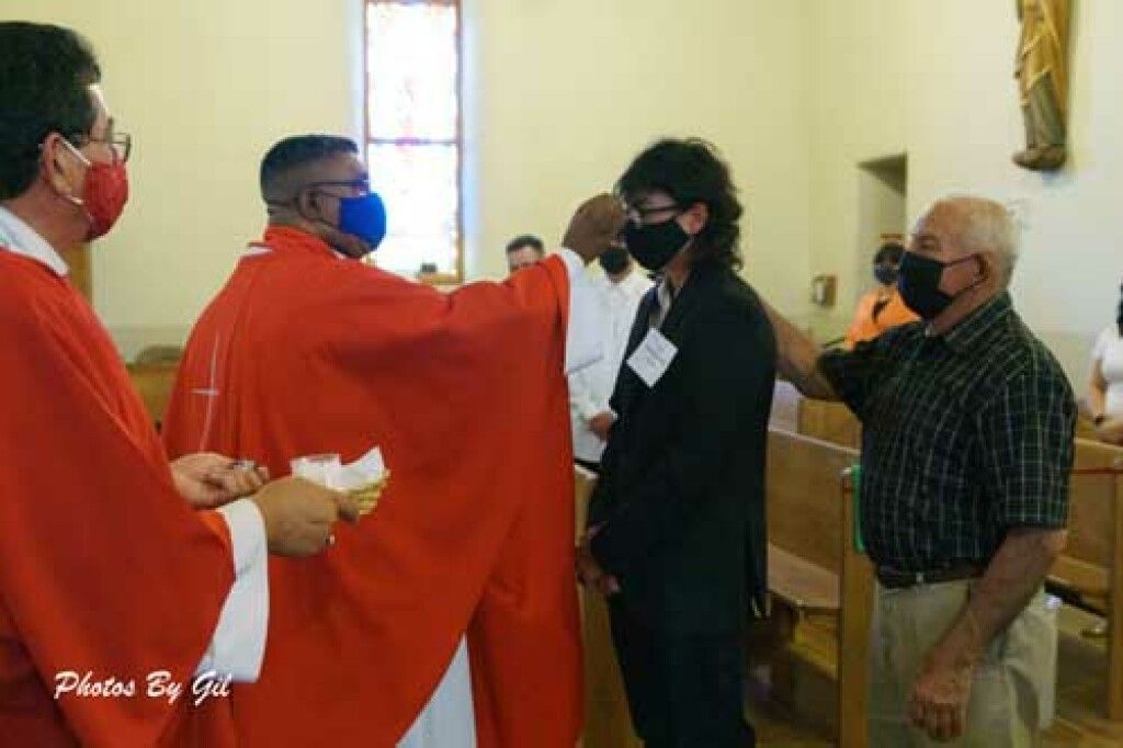 A priest in red robes performs a religious blessing on a masked young man inside a church.