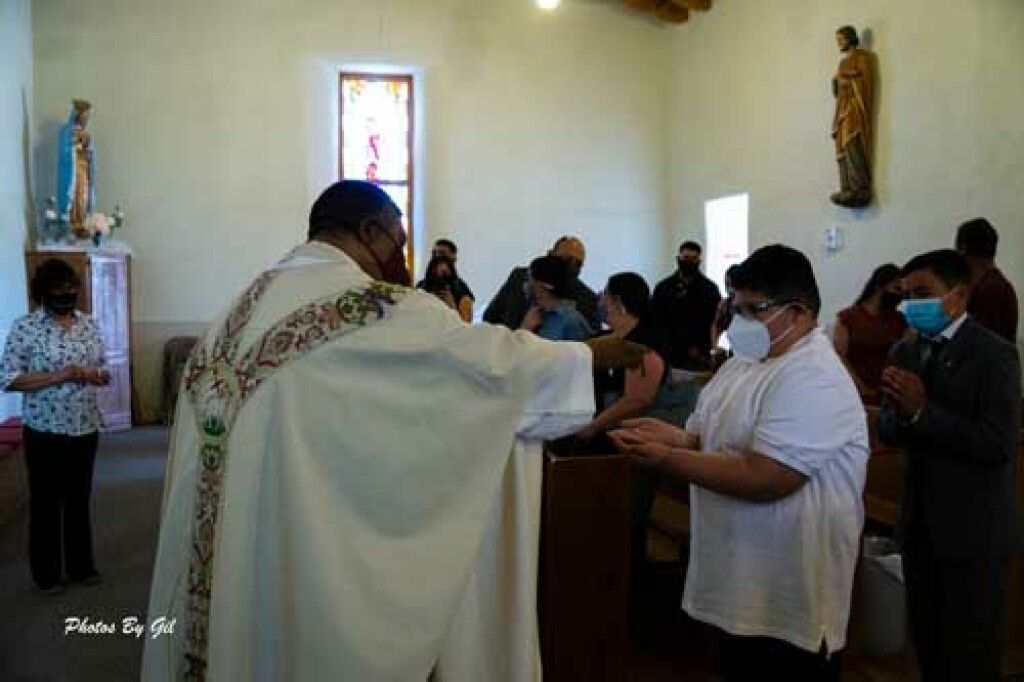 A priest in a white robe blesses a masked congregation in a church. 