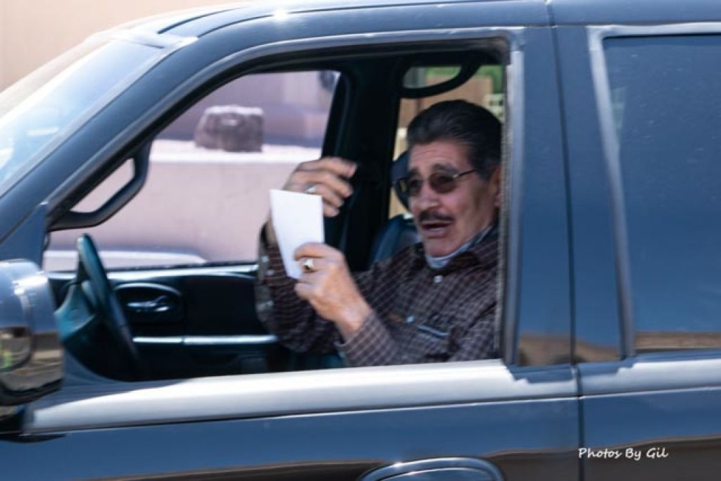 A man in sunglasses and a brown shirt is in a car, holding a piece of paper.