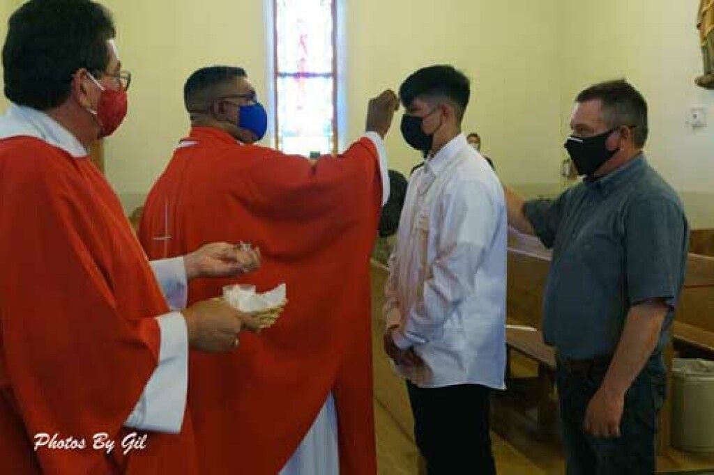 A priest in red robes anoints a young man wearing a white shirt and black mask in a church.