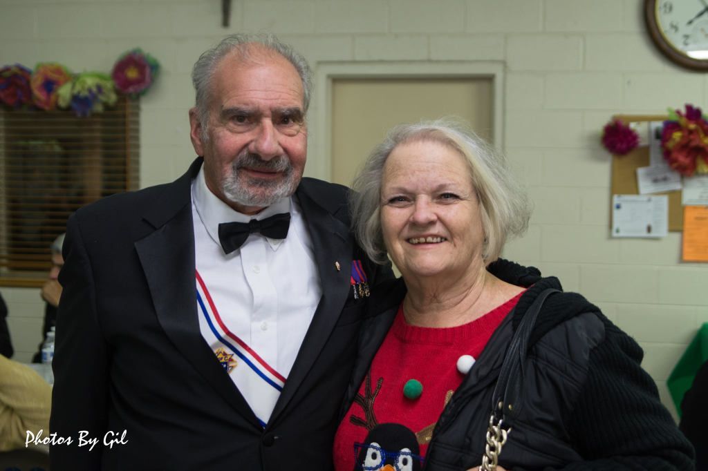 Elderly man in a tuxedo with medals, and woman in a festive red sweater.