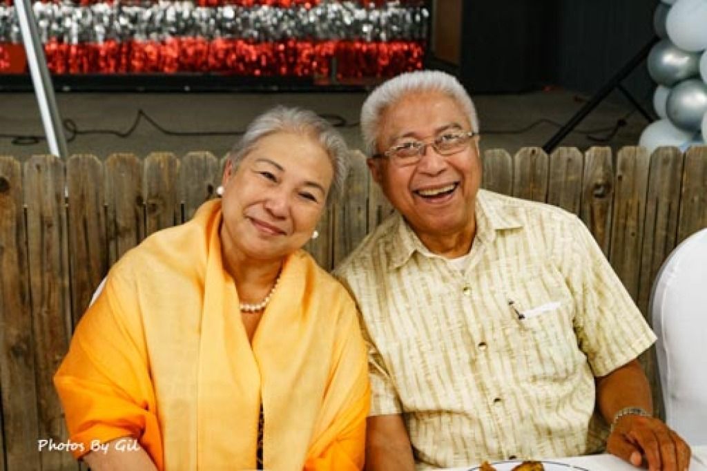 Elderly couple smiling warmly at a party, sitting close. 