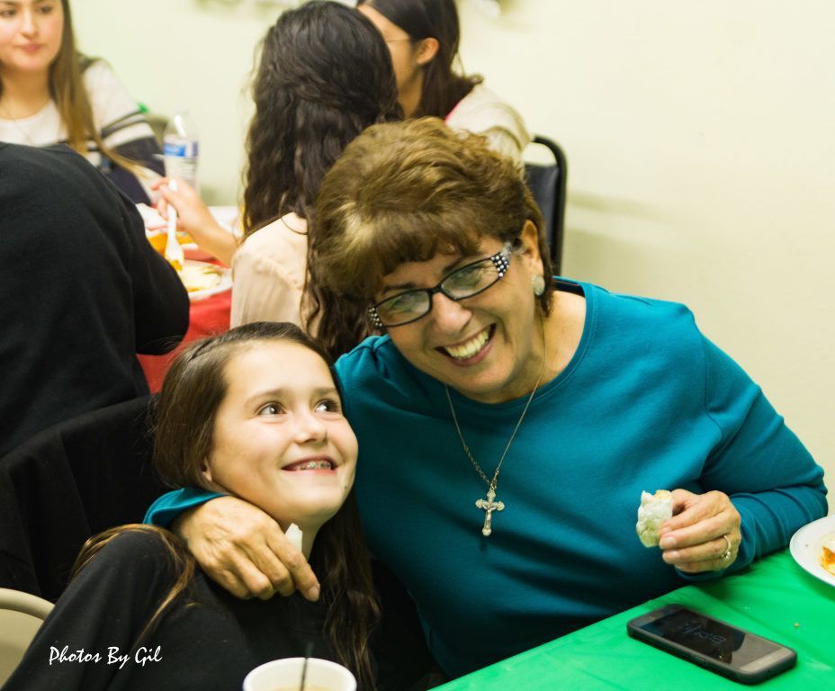 A smiling woman in glasses and turquoise top hugs a young girl at a table set for a meal.