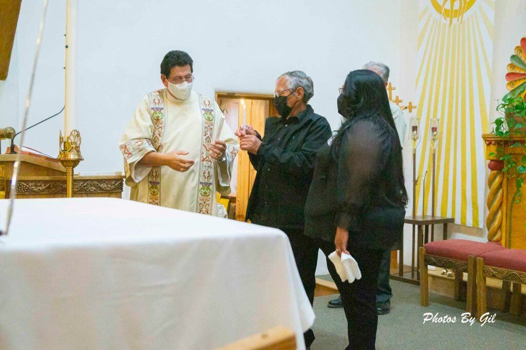 A priest in ornate robes shares a lit candle with two masked individuals in a church.
