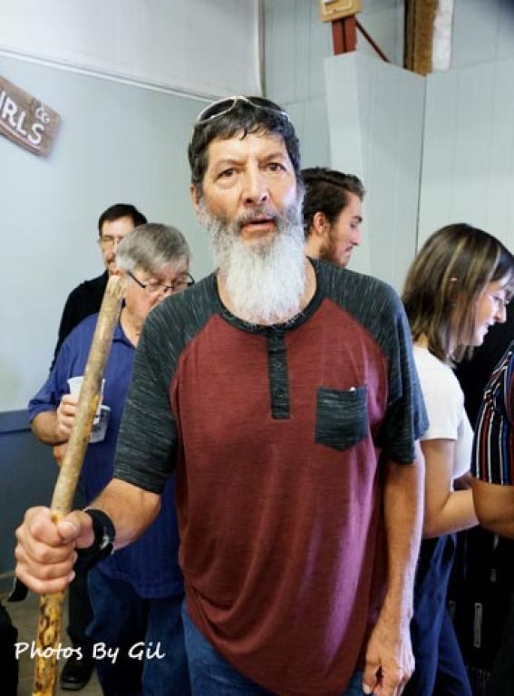 A bearded man with a walking stick stands in a room with several people behind him.