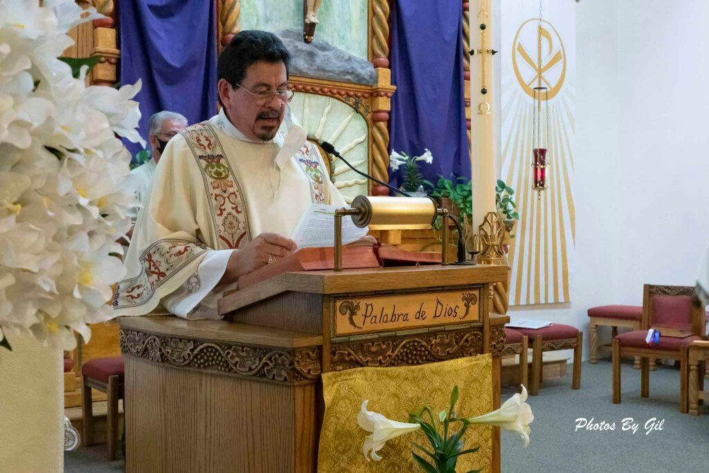 A priest stands at a wooden pulpit reading, flanked by white lilies.