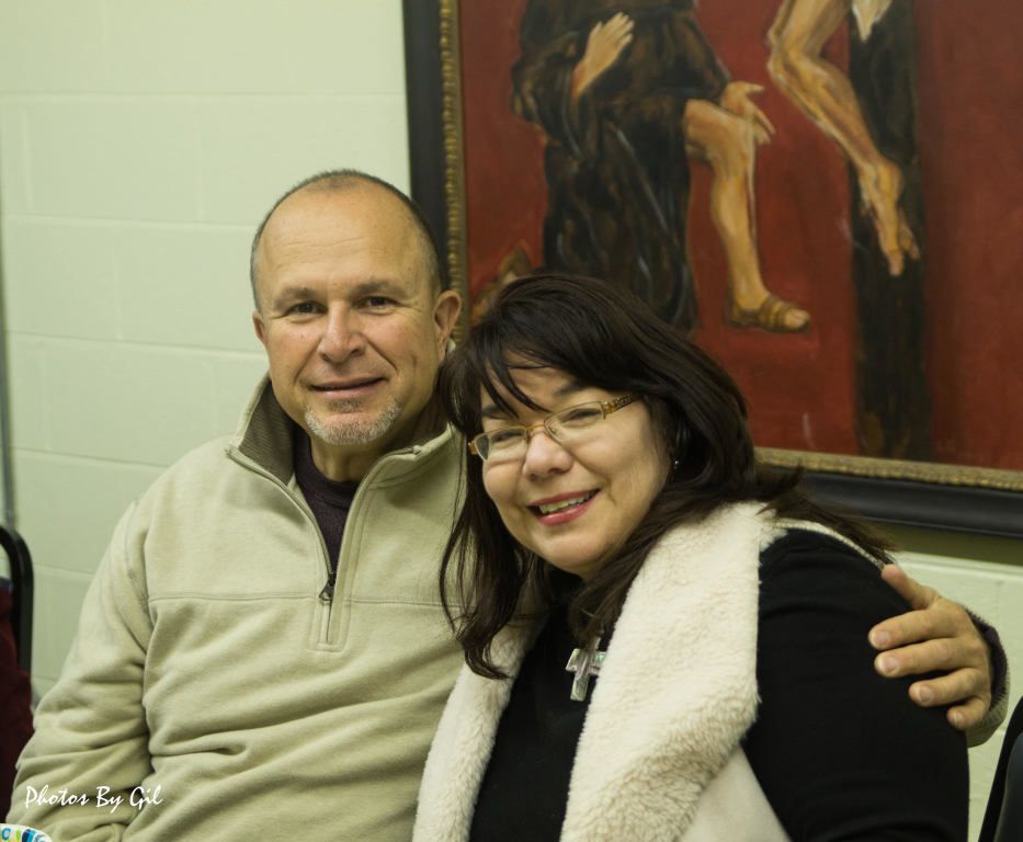 A smiling couple sits closely in a warmly lit room, with a large painting.