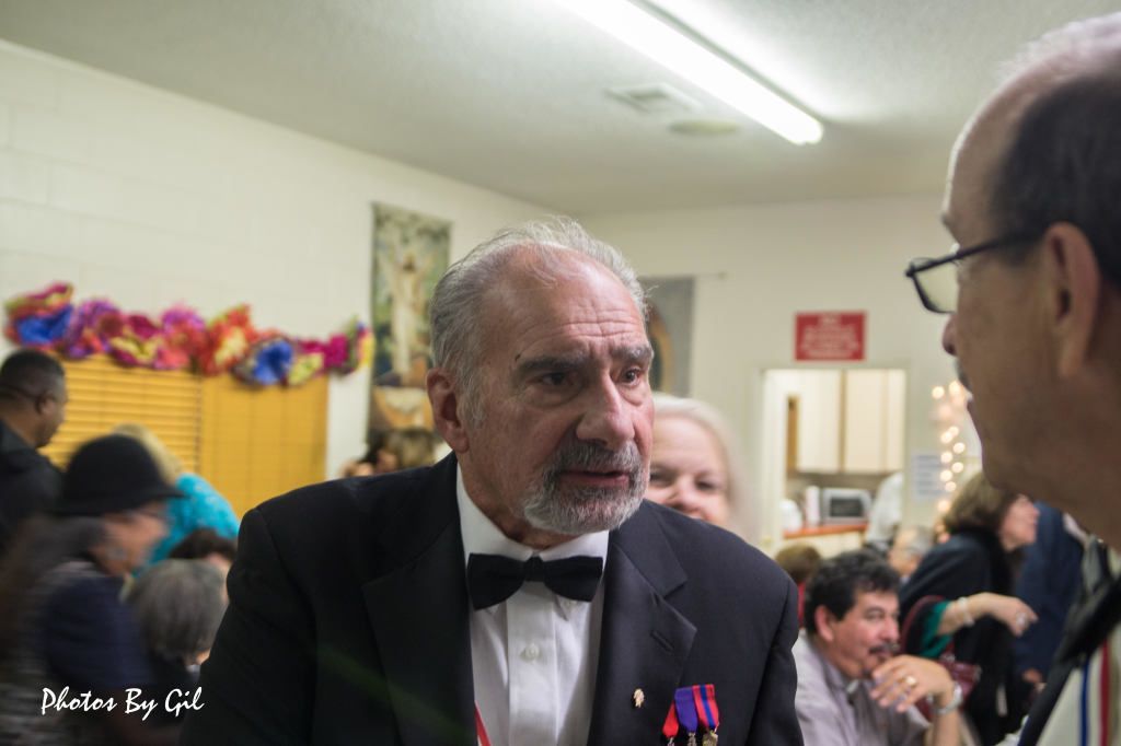 An elderly man in a tuxedo, with medals on his chest, is talking to another man in glasses.