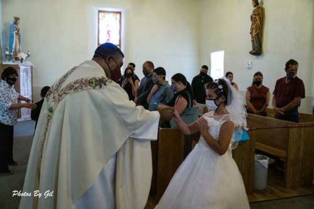 A priest and a girl in a white dress, both wearing masks, share a handshake during a church ceremony. 